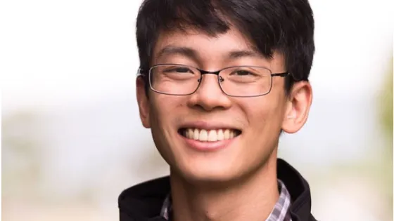 A smiling young man with glasses wearing a black jacket and plaid shirt.