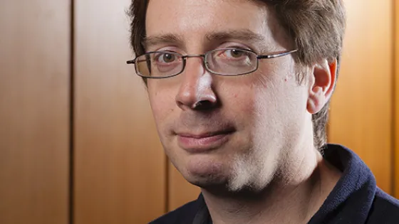 A portrait of a man with light brown hair and glasses, wearing a dark jacket, standing in front of a wooden paneled background.