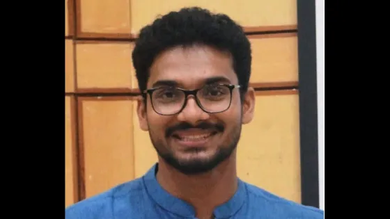 A portrait of a young man with glasses, smiling, wearing a blue shirt against a background of beige tiles.