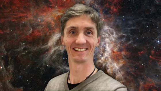 A man with a friendly smile, wearing a grey shirt, standing against a vibrant cosmic starry background.