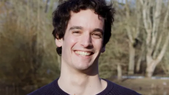 A young man smiling in a park with trees and a pond in the background.