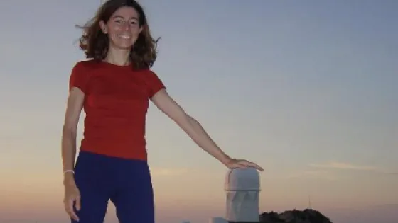 A smiling woman in a red shirt stands atop a hill at dusk, with several large telescope observatories in the background.