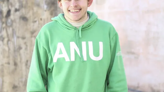 A young man wearing a green hoodie and a cap smiles in front of a textured wall.