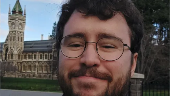 A man with glasses smiling in front of a historic building with a clock tower.