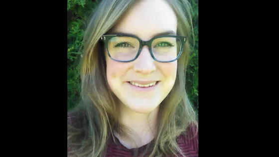 A smiling woman with glasses and long hair, standing in front of a green, leafy background.
