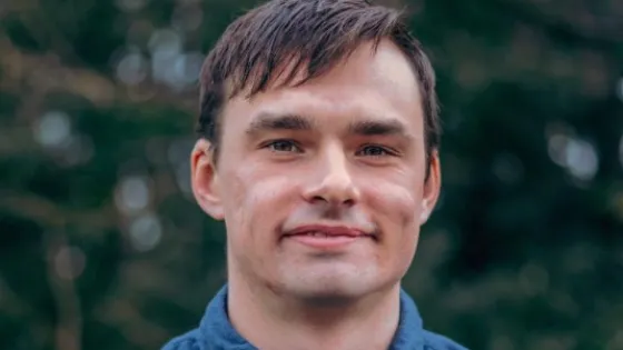 A portrait of a young man with short brown hair, smiling, wearing a blue shirt, with a blurred green background.