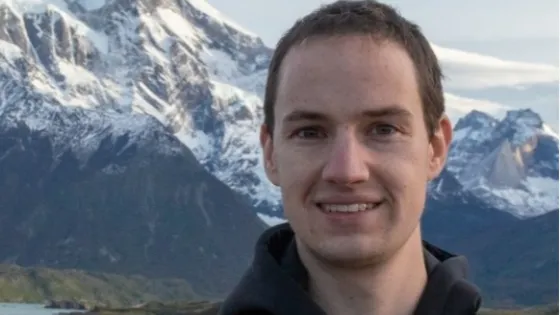 A man smiling at the camera with snowy mountains and a lake in the background.