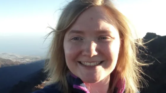 A smiling woman with blonde hair takes a selfie with mountainous scenery visible in the background under a clear sky.