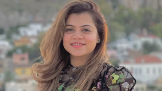 A woman with long, wavy hair smiles at the camera, standing before a blurred background of a town nestled in a rocky landscape.