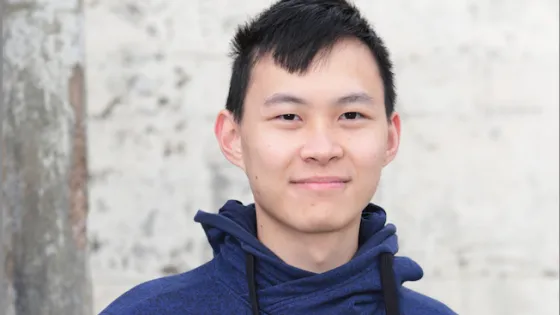 A young man in a blue hoodie smiling at the camera with a neutral background.