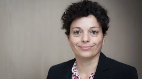 A professional portrait of a woman with curly hair wearing a blazer and a patterned blouse, smiling slightly against a neutral background.