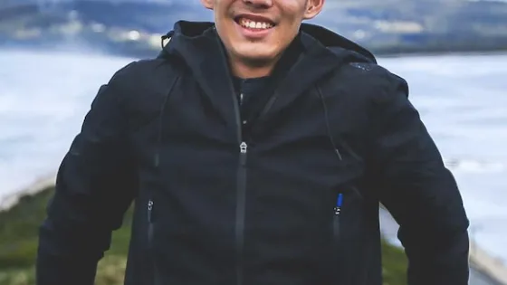 A smiling man wearing glasses and a black jacket stands by a railing with a blurred landscape in the background.