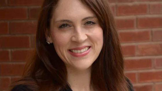 A woman with long brown hair smiling in front of a brick wall.