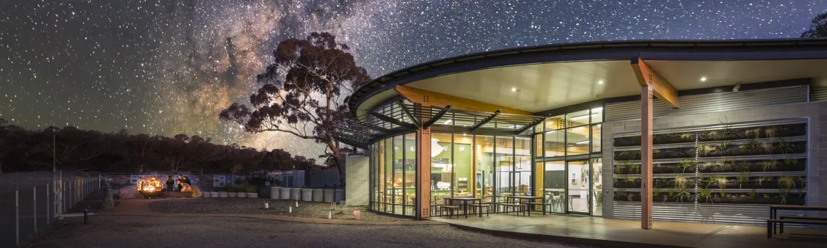 A modern building with glass walls under a star-filled sky, surrounded by trees and a small group of people gathered around a campfire nearby.