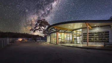 A modern building with glass walls under a star-filled sky, surrounded by trees and a small group of people gathered around a campfire nearby.