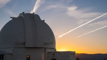 A large astronomical observatory dome captured at sunset with long streaks in the sky created by airplane contrails.