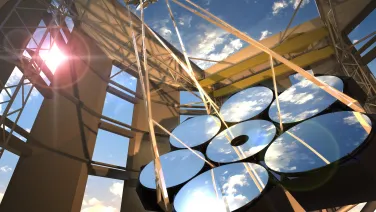 A large telescope under a bright sun with its mirrors reflecting the blue sky and clouds, set against a steel structure.
